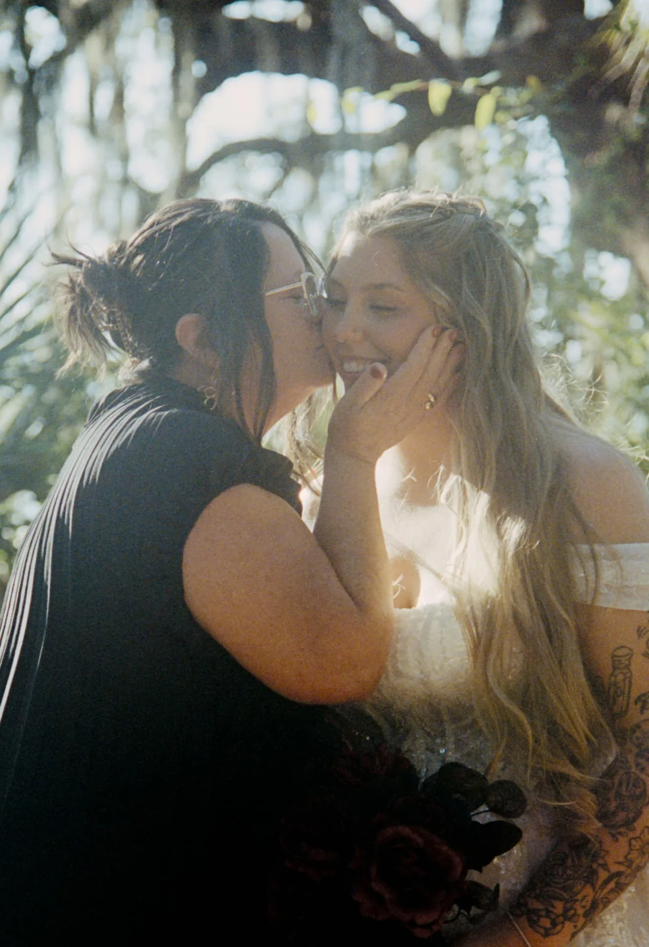 Mother kissing the bride's cheek as she holds a burgundy bouquet beneath sunlit oaks, photographed on Portra 400 wedding film