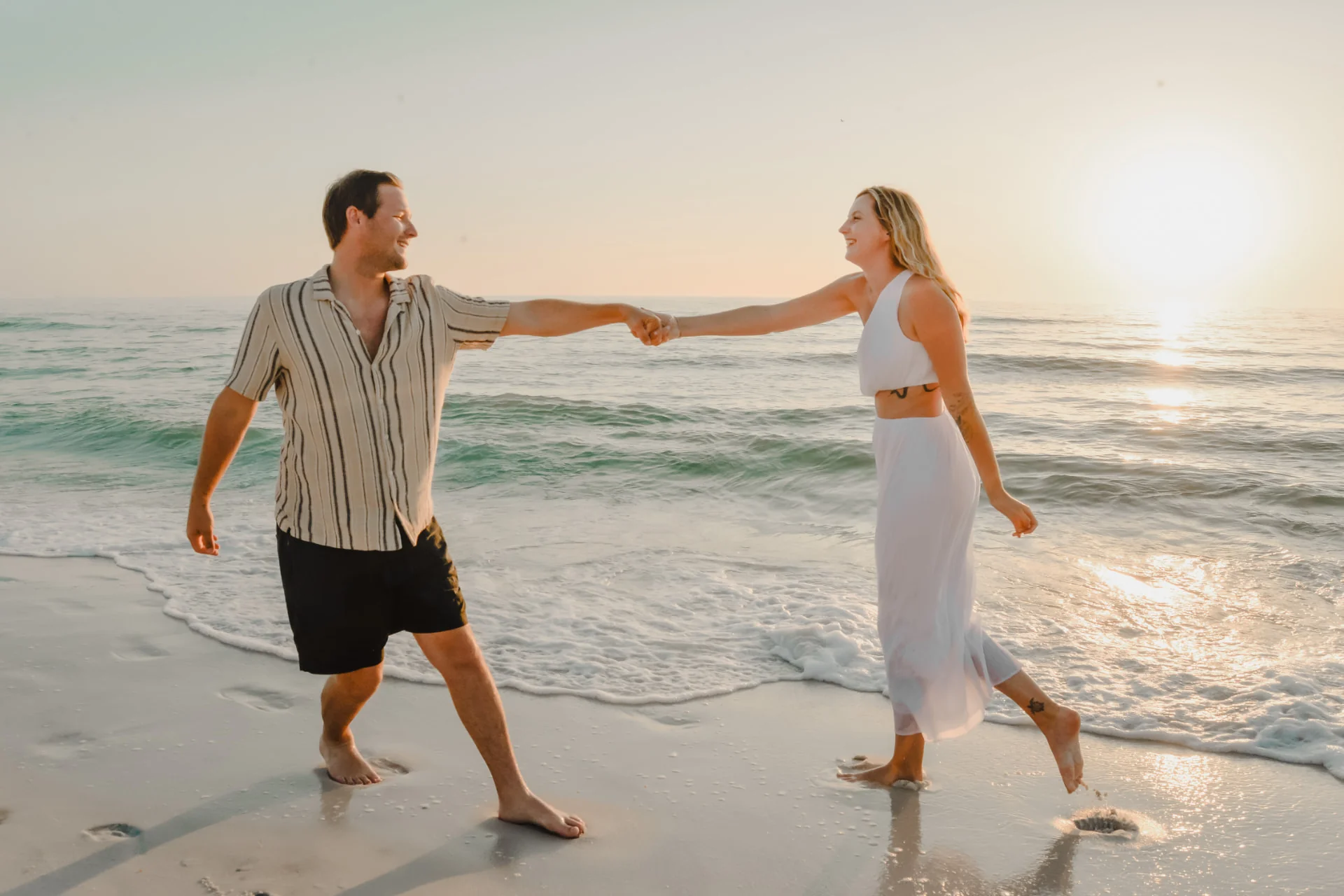 Engaged couple holding hands while walking along the shoreline at sunset