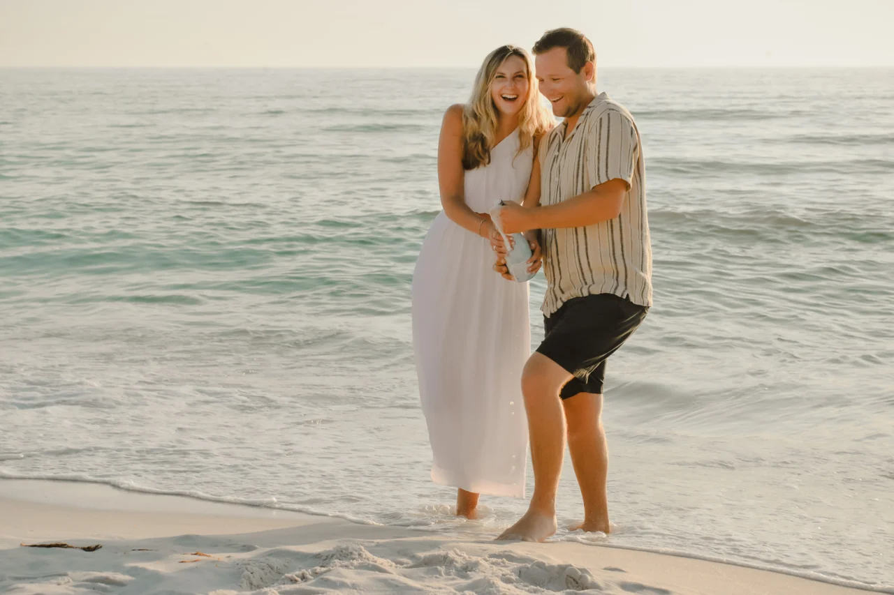 Engaged couple laughing while opening champagne in the surf at sunset
