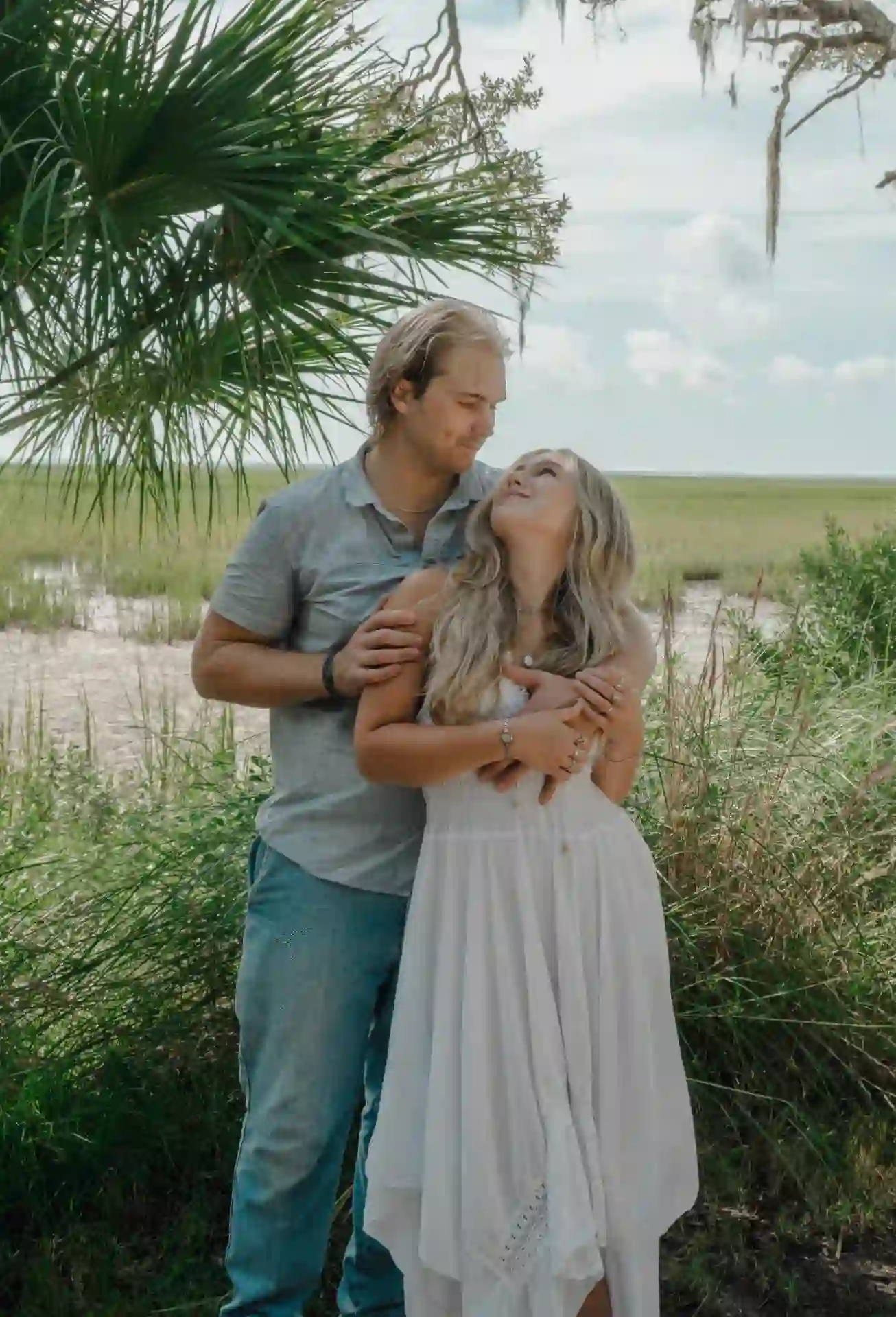 Engaged couple holding each other under Spanish moss beside a tidal marsh