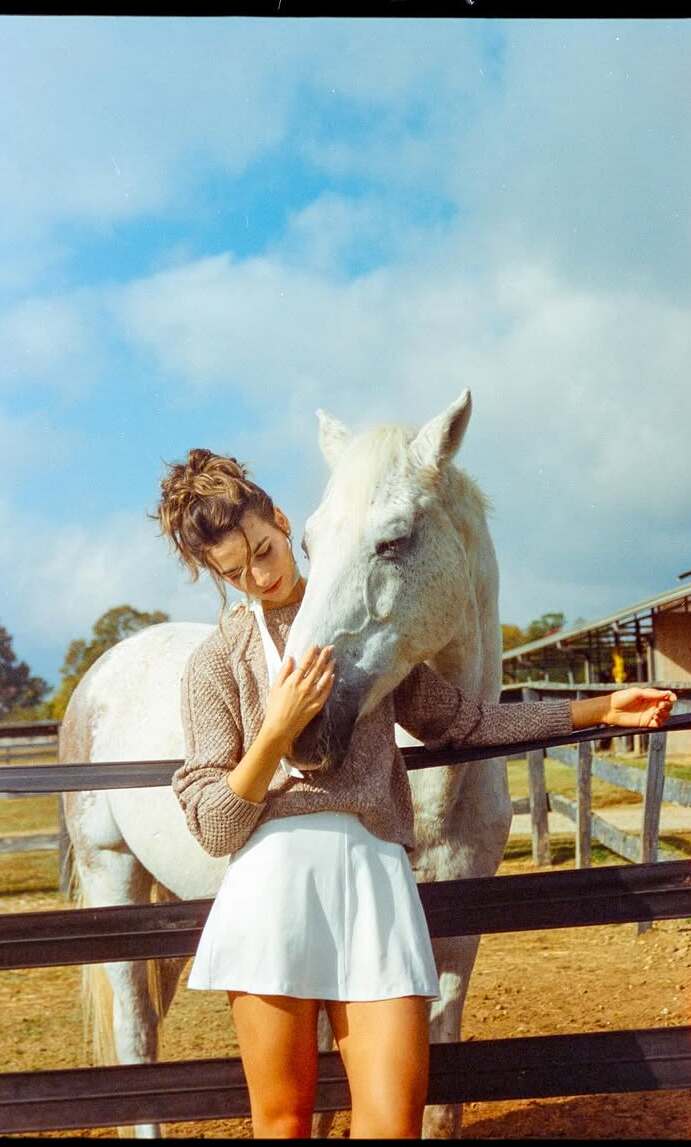 Editorial portrait in a white cardigan beside a horse with soft film color