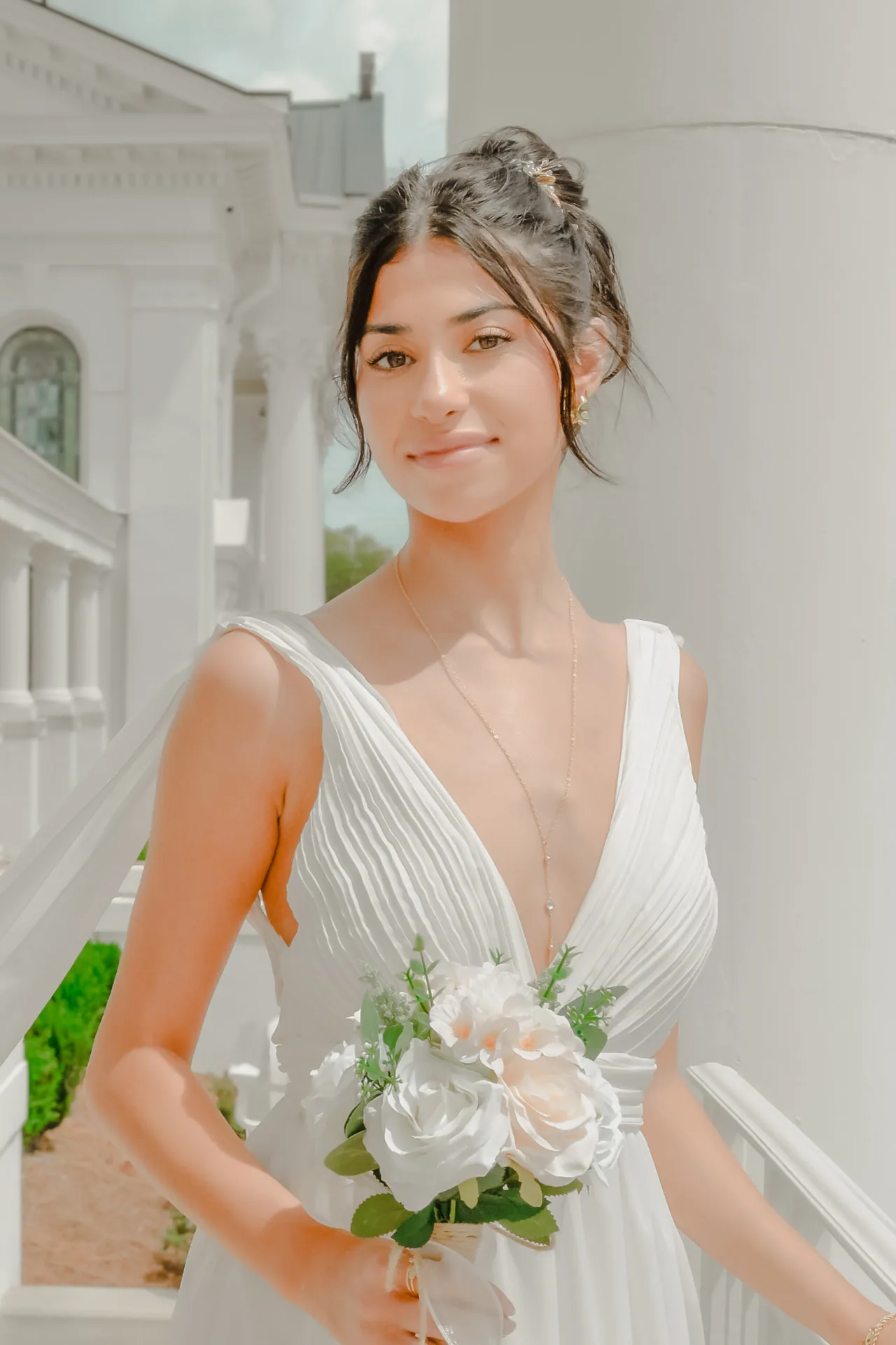 Bride in a pleated white gown holding a bouquet on a sunlit porch