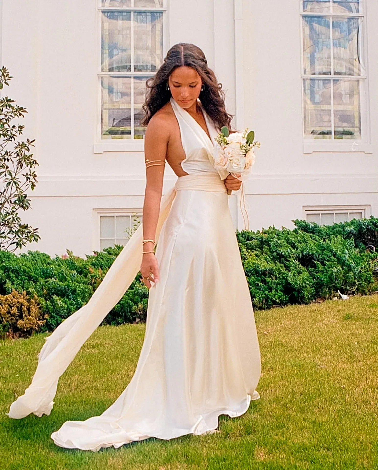 Bride in a white halter dress holding a bouquet on a green lawn