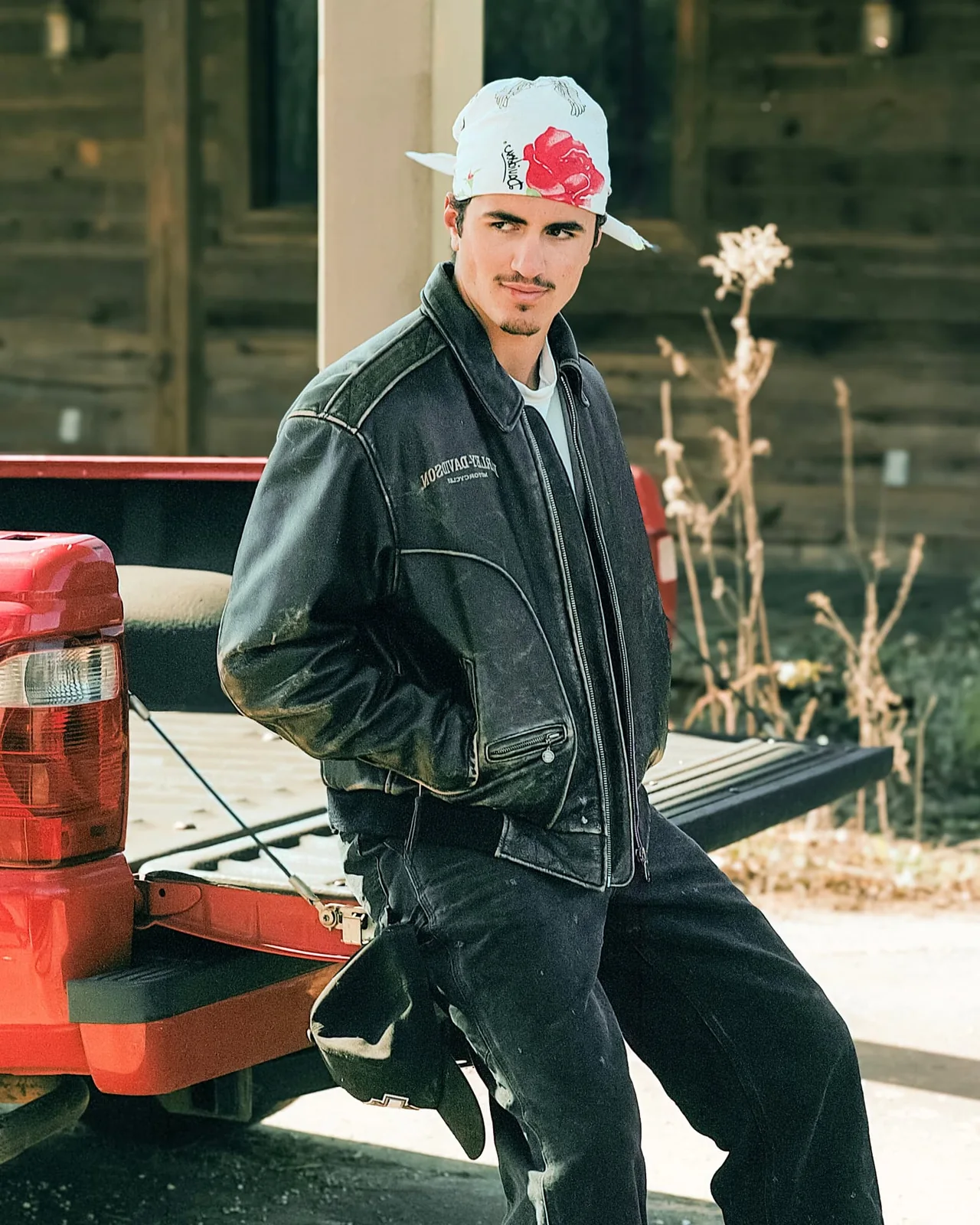 Editorial portrait in a leather jacket and floral bandana beside a red pickup truck