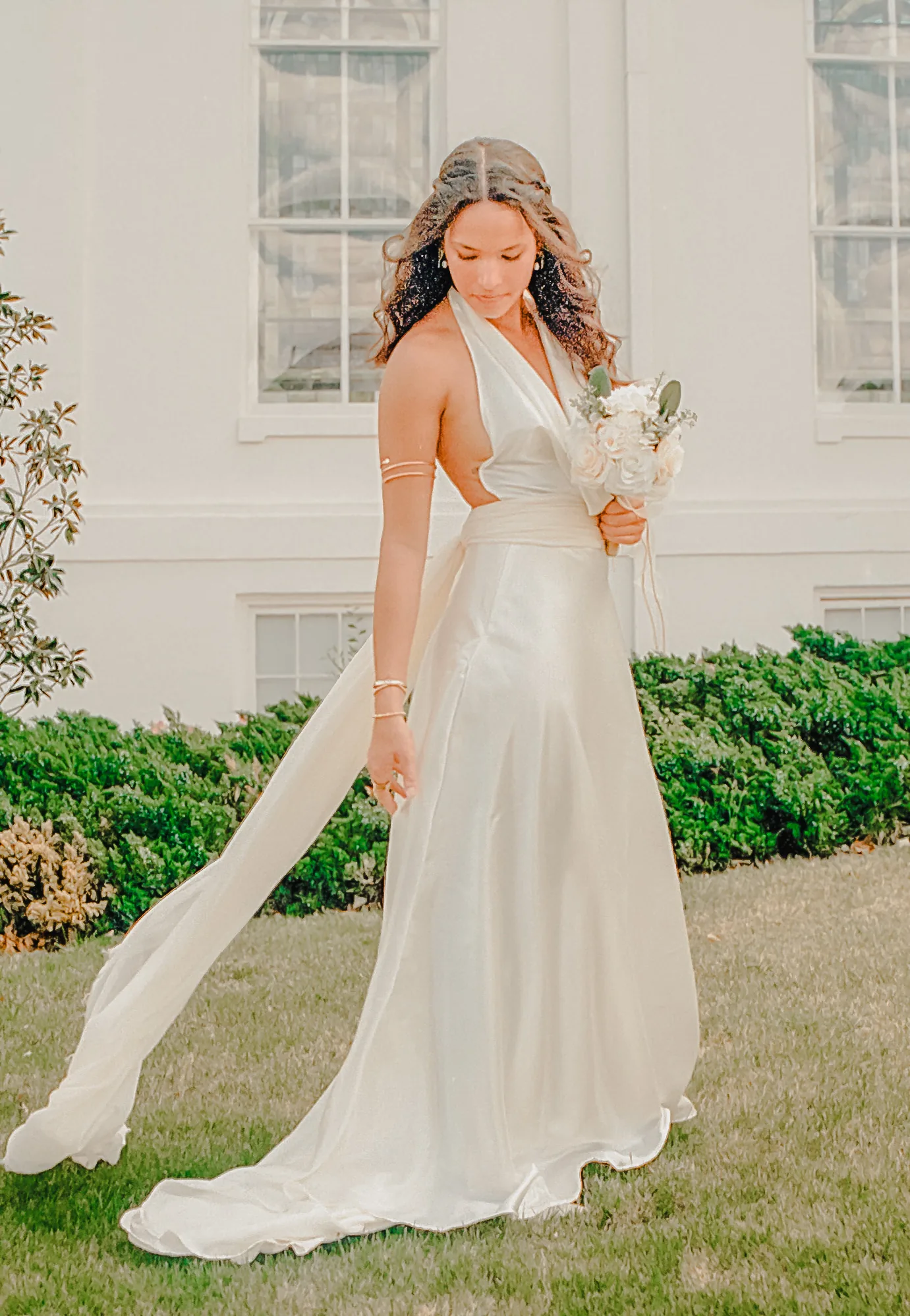 Bride in a white halter dress holding a bouquet on a green lawn