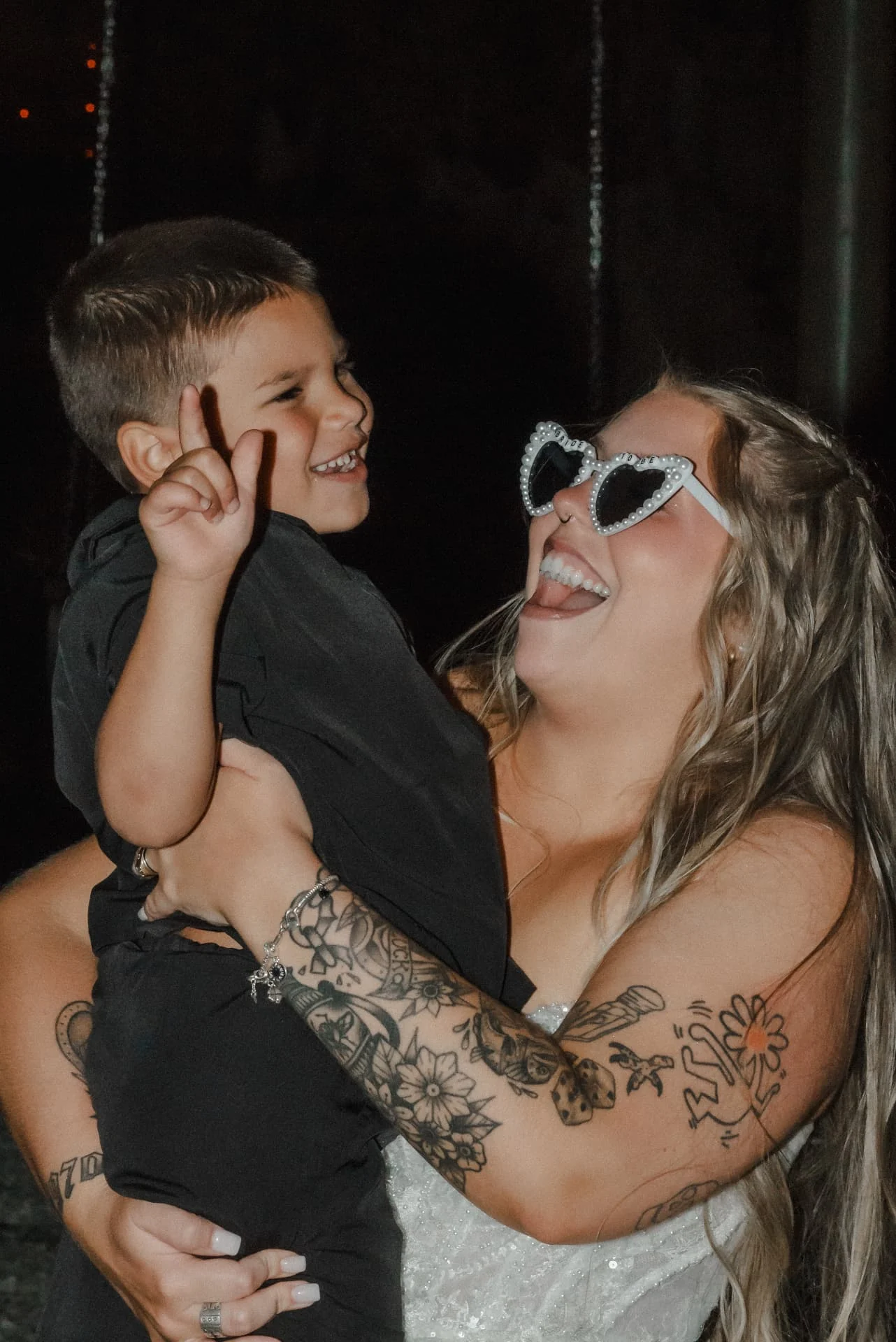 Bride wearing heart-shaped sunglasses and laughing with a young boy at the reception