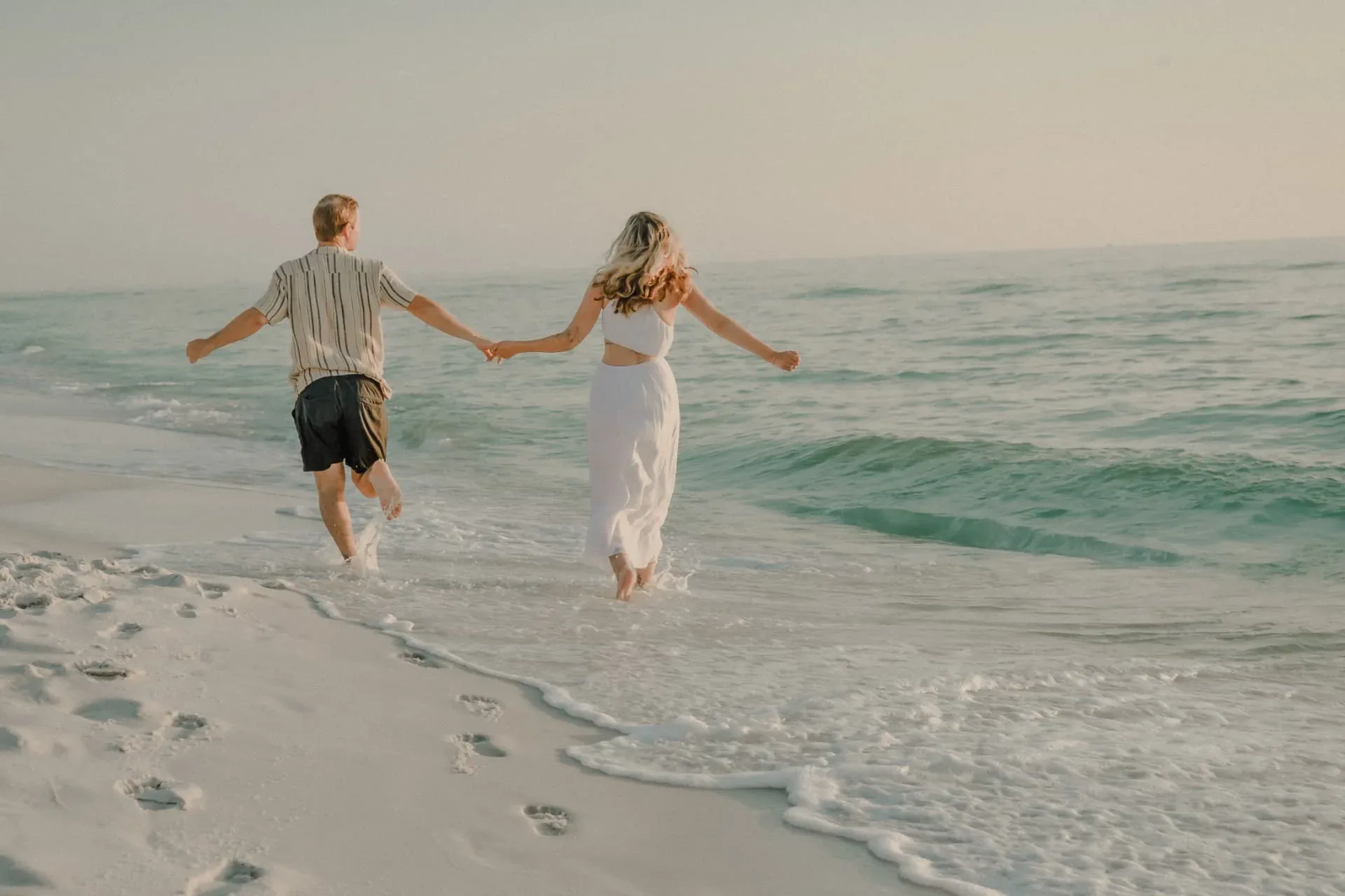 Engaged couple running hand in hand into the surf at golden hour