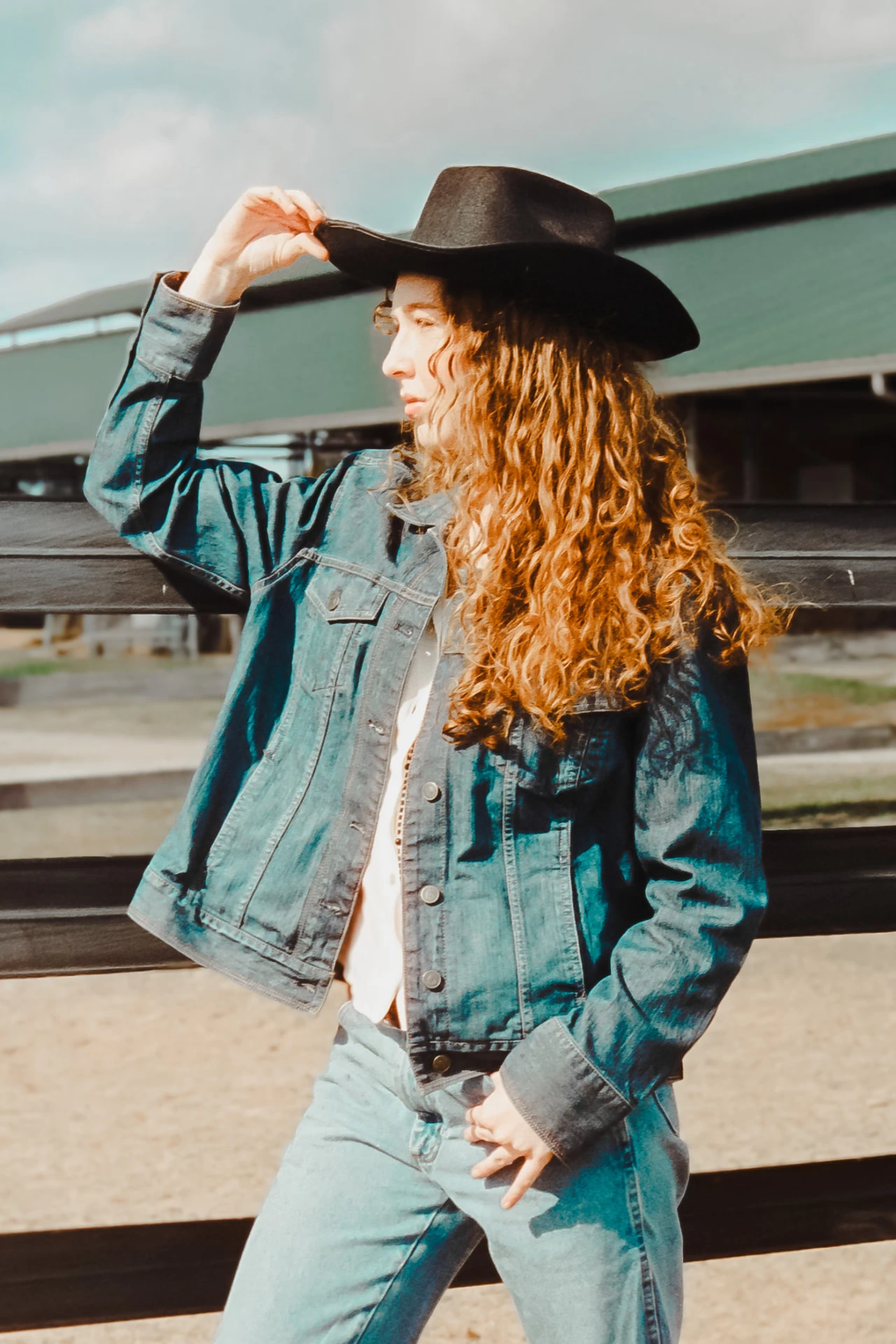 Editorial portrait of a red-haired woman in a black cowboy hat and denim jacket leaning against a ranch fence on CineStill 400D film