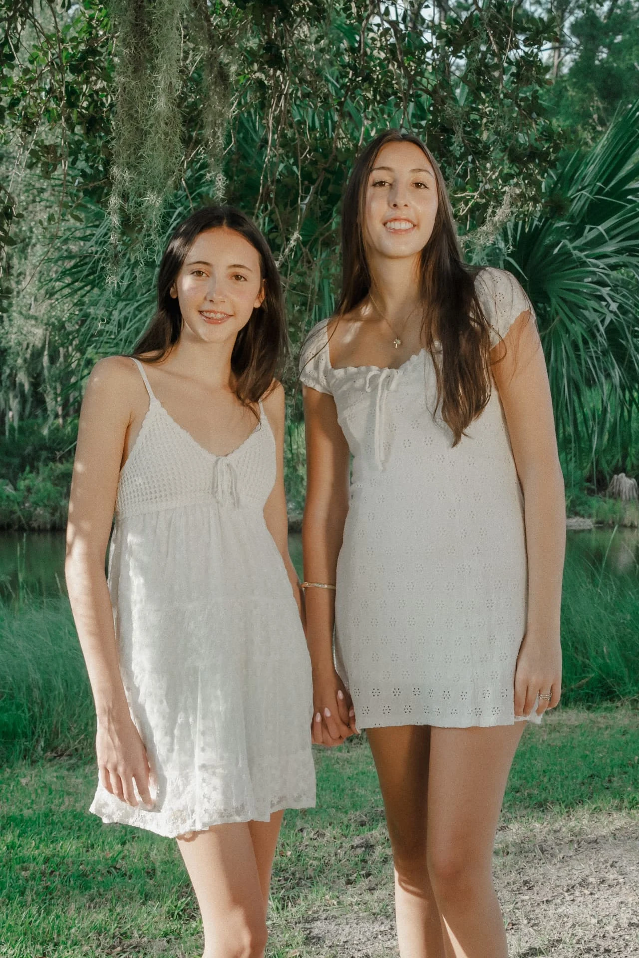 Two sisters holding hands in white dresses beneath the trees